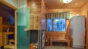 Interior de uma cabana de madeira, com um banco, uma geladeira, uma janela com vista para árvores e uma porta com painel de vidro fosco.