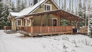 Uma cabana de tronco com varanda de madeira e chaminé, rodeada por árvores cobertas de neve.