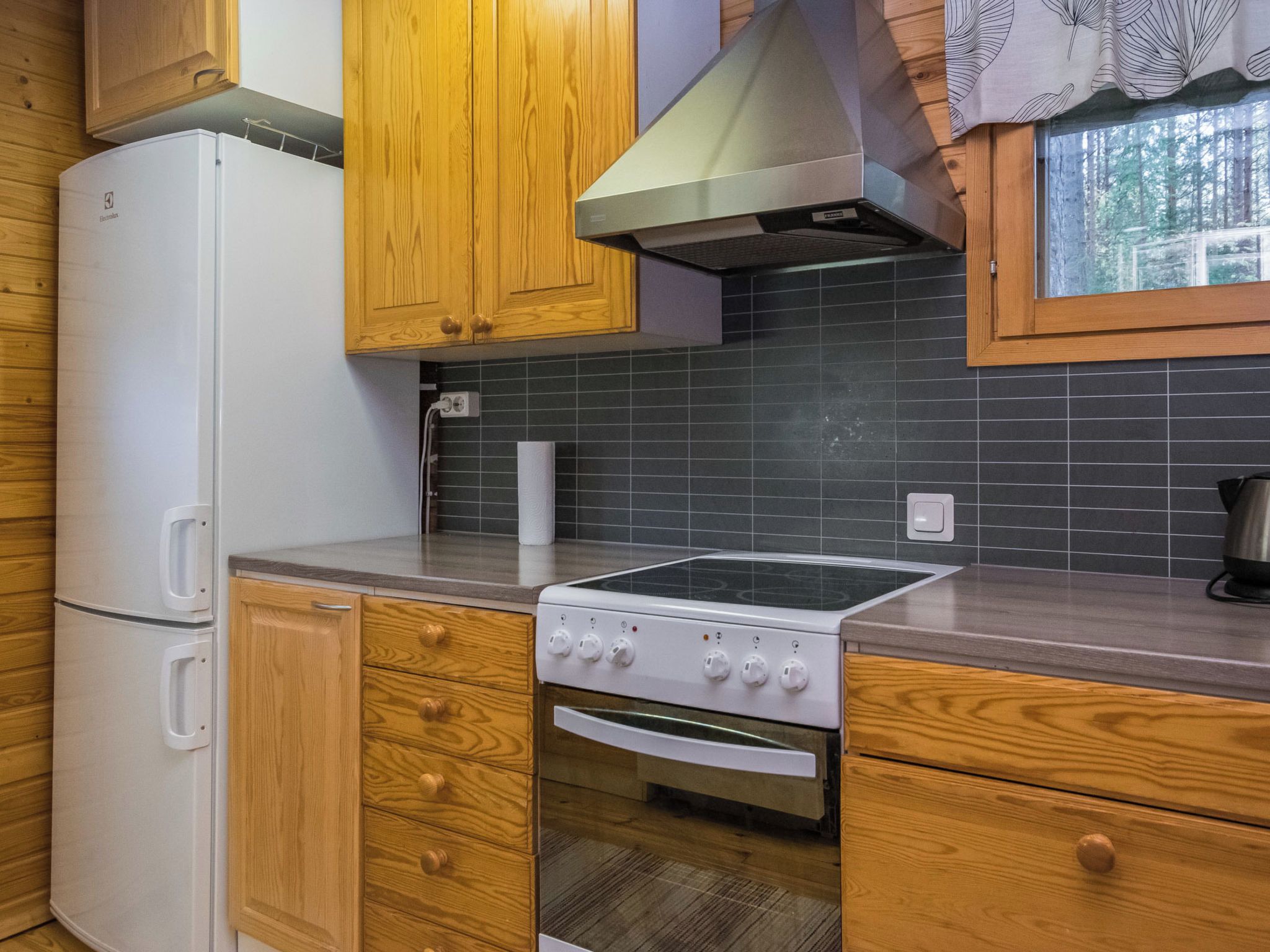 Une cuisine avec des meubles en bois, un réfrigérateur blanc, une cuisinière et une fenêtre donnant sur des arbres.