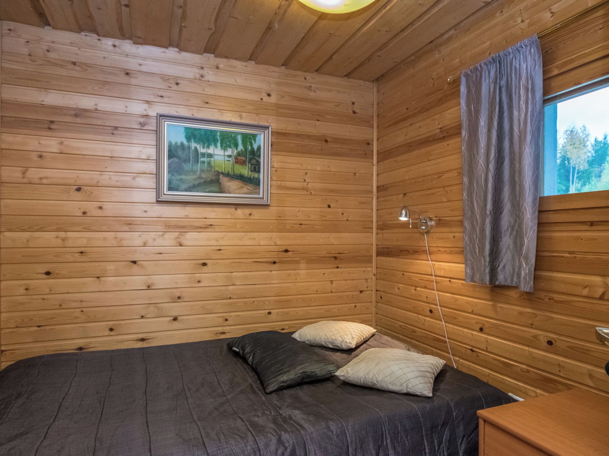 A wooden cabin room with a bed, pillows, a framed picture, a lamp, and a window with curtains.