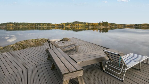 A wooden deck with picnic tables and a lounge chair overlooking a calm lake with autumn foliage in the background.