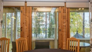A dining area with a round table and four wooden chairs, large windows with wooden frames, and a view of trees and a building outside.
