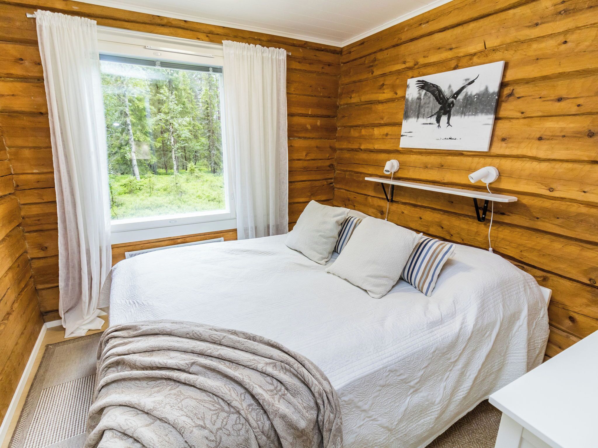 Quarto de cabana de madeira com cama, janela com cortinas, um quadro e uma mesinha de cabeceira.