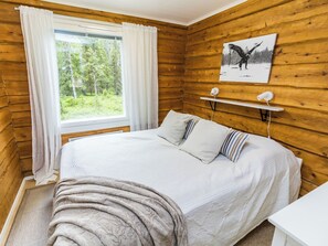 A wooden cabin bedroom with a bed, a window with curtains, a painting, and a bedside table.