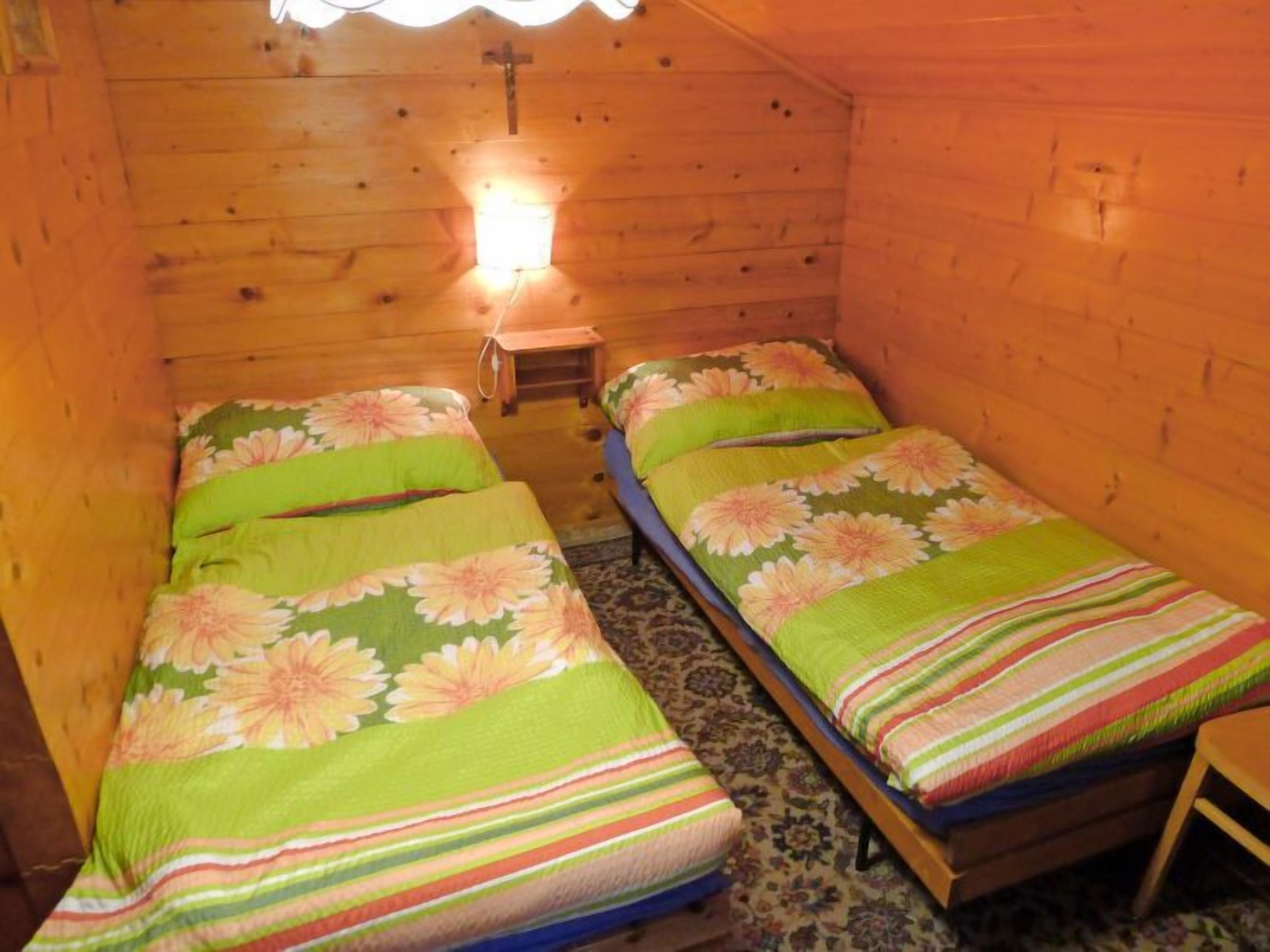 Eine kleine Holzhütte mit zwei Einzelbetten, jeweils mit blumigem Bettbezug, einem kleinen Tisch und einem Kruzifix an der Wand.