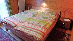 A wooden cabin bedroom with a bed, a nightstand, a radiator, and a window with curtains. - Lärchenheim by Interhome (St Niklaus)