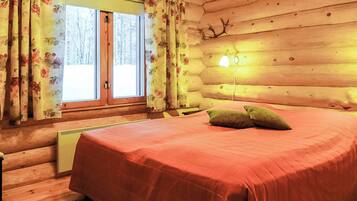 A log cabin bedroom with a bed, curtains, a lamp, and a view of a snowy landscape.