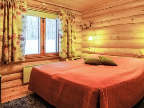 A log cabin bedroom with a bed, curtains, a lamp, and a view of a snowy landscape.