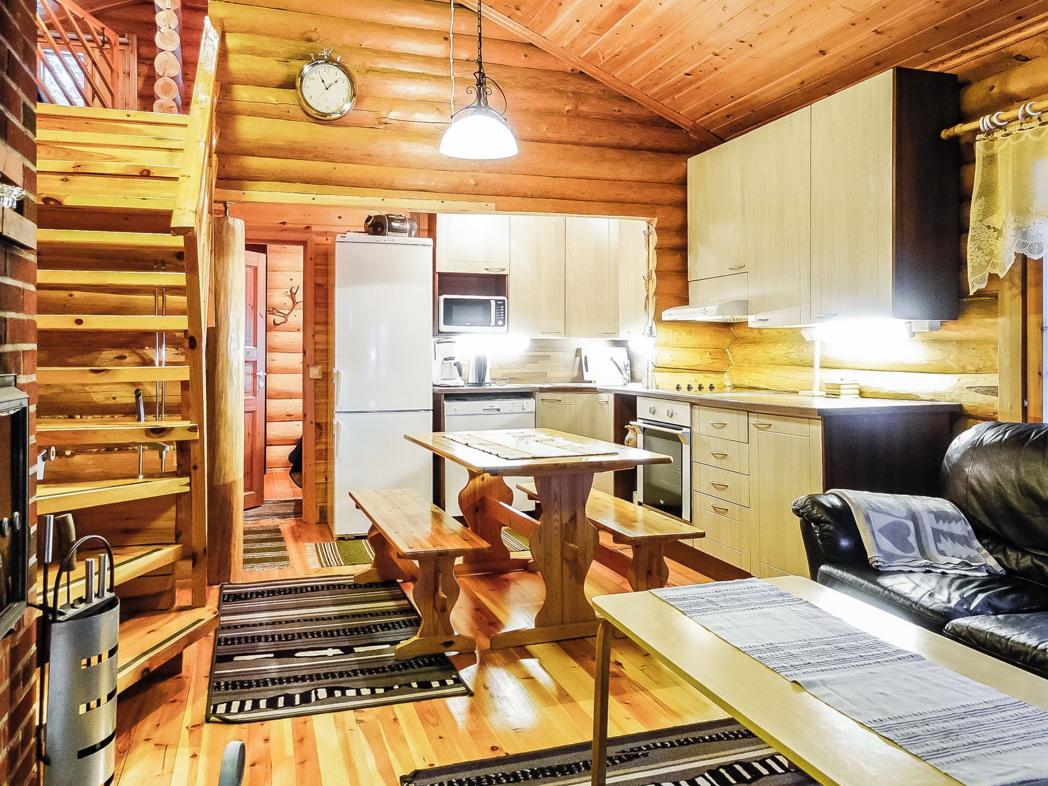 A log cabin interior with a wooden dining table, benches, a kitchen area with a microwave, and a fireplace.