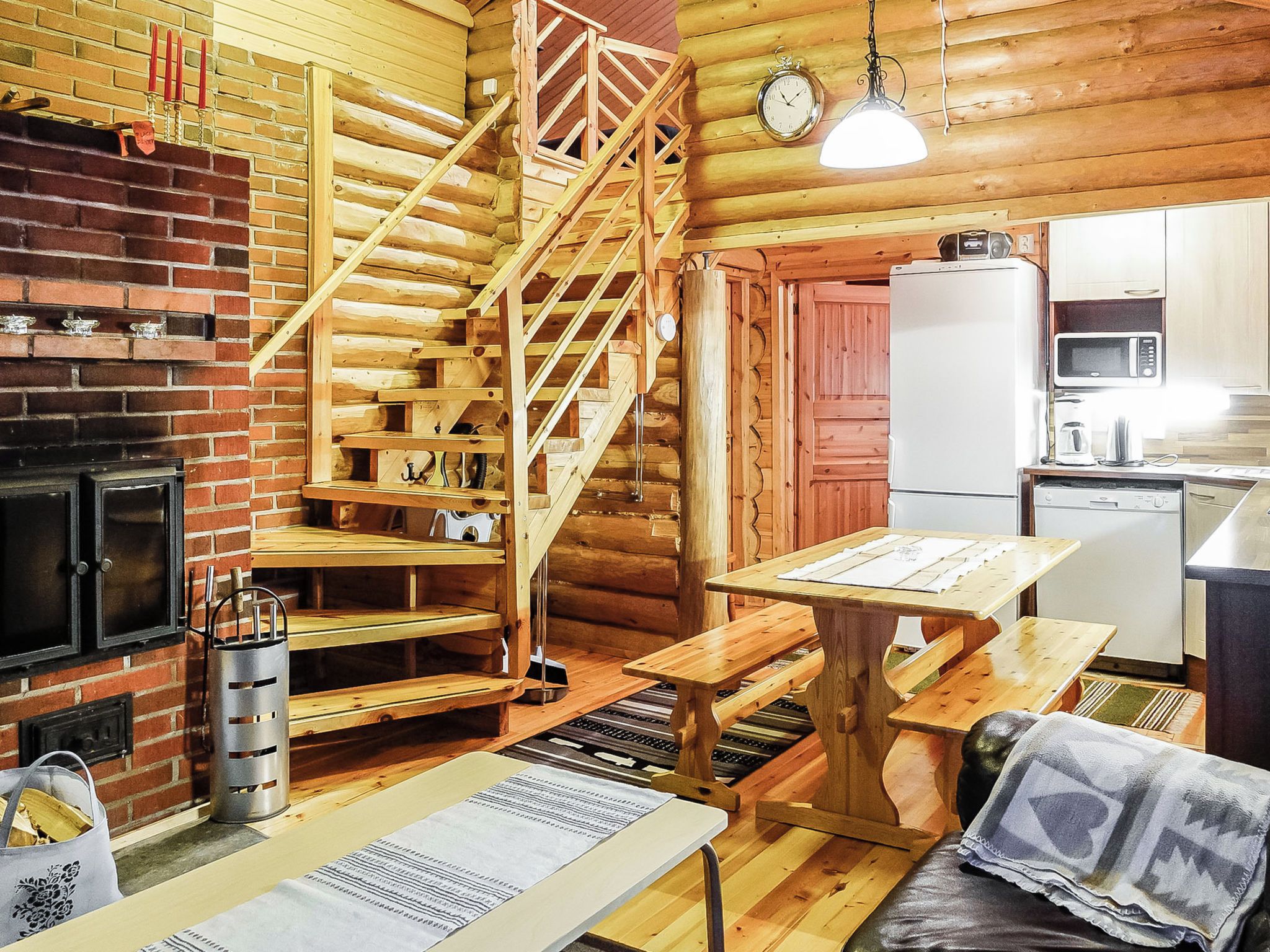 A log cabin interior with a brick fireplace, wooden staircase, a dining table, and a kitchen area.