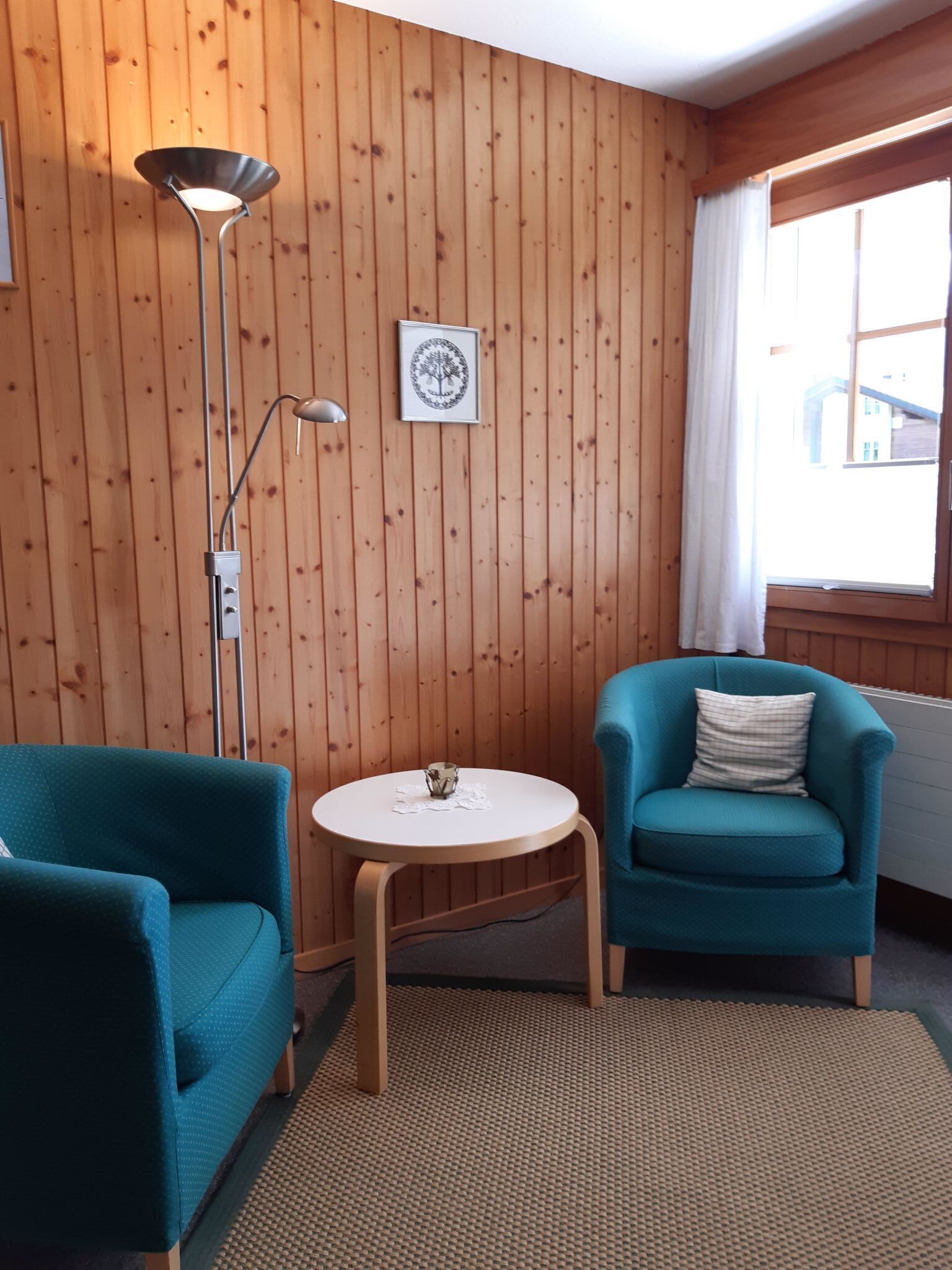A room with wooden panel walls, a round table with a white tablecloth, two teal armchairs, a floor lamp, and a framed picture on the wall.