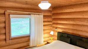 A wooden cabin room with a bed, a window with curtains, a nightstand with a lamp, and a view of a grassy field. - Suviranta by Interhome (Lapinlahti)