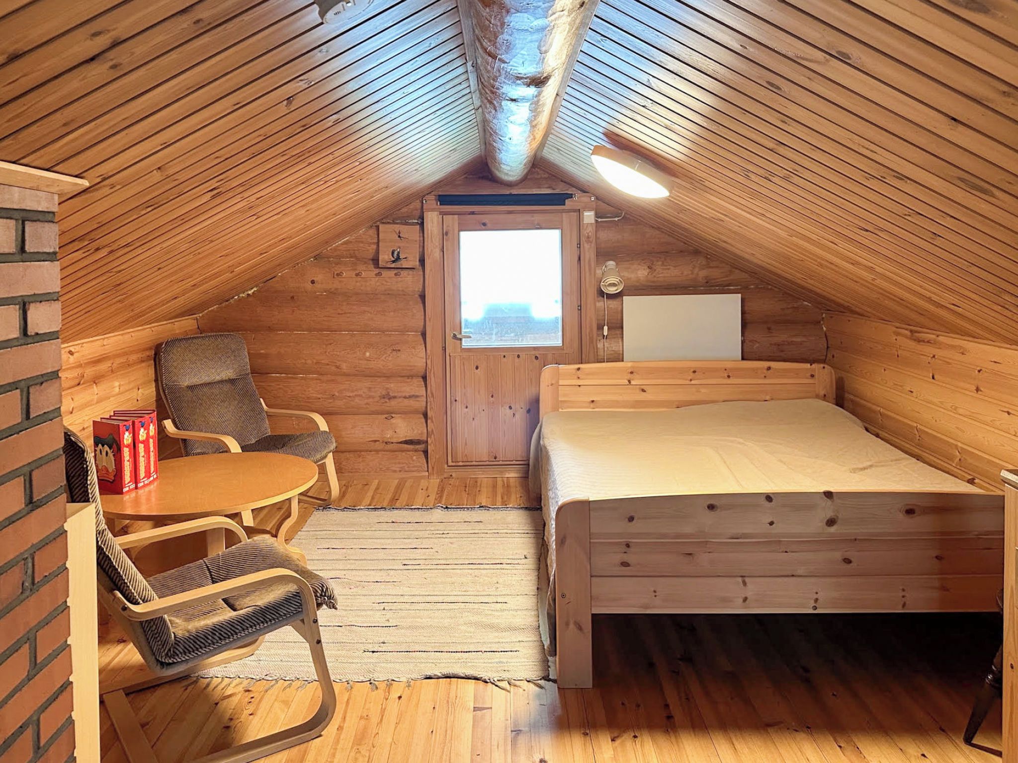 Un dormitorio acogedor en el ático con techo inclinado de madera, una cama individual, una mesita, dos sillas y una ventana.