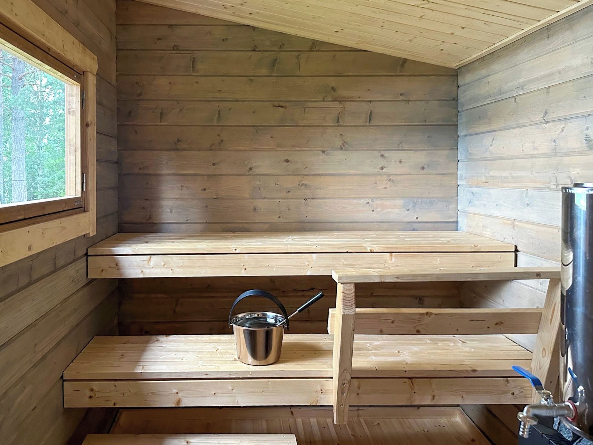 L’intérieur d’une cabane en bois, avec une fenêtre, un seau en métal et un robinet.
