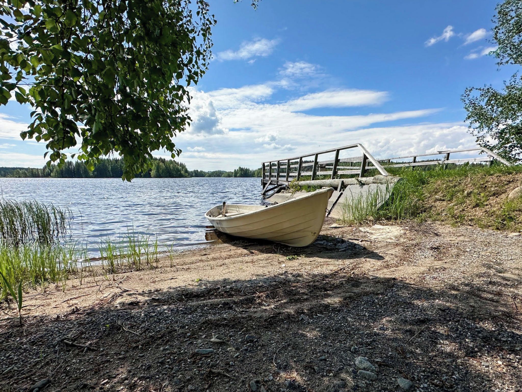Un bateau sur une plage de sable, avec une jetée en bois qui s’avance dans un lac, le tout entouré d’arbres et sous un ciel dégagé.