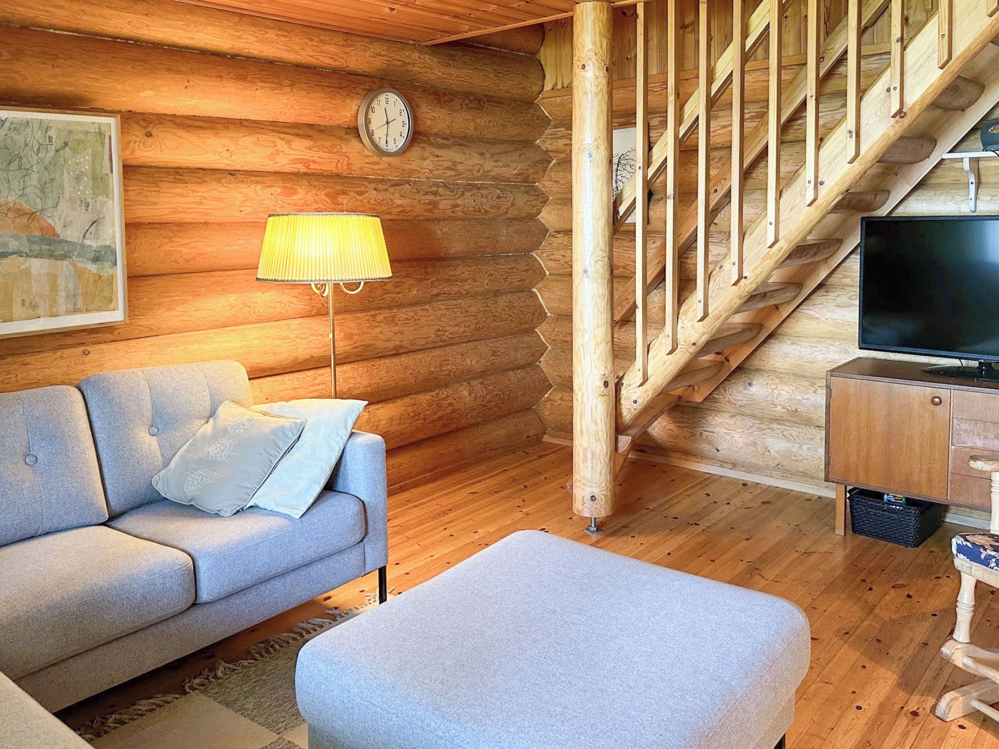 L’intérieur d’une cabane en rondins, avec un canapé, un pouf, une lampe, un téléviseur et un escalier en bois.