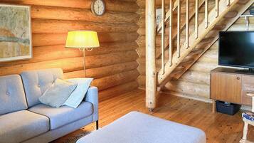 L’intérieur d’une cabane en rondins, avec un canapé, un pouf, une lampe, un téléviseur et un escalier en bois.