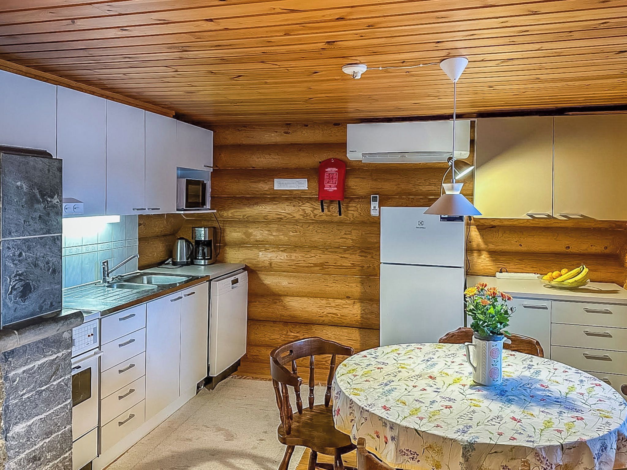 A kitchen with wooden walls, white cabinets, a microwave, a coffee maker, a refrigerator, and a dining table with a floral tablecloth.