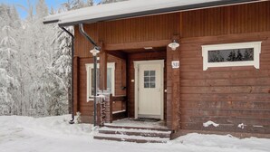 A wooden cabin with a front door, a staircase leading up to the entrance, and a snow-covered landscape with trees. - Klz 11. jade by Interhome (Sotkamo)
