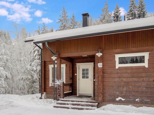 A wooden cabin with a front door, a staircase leading up to the entrance, and a snow-covered landscape with trees.