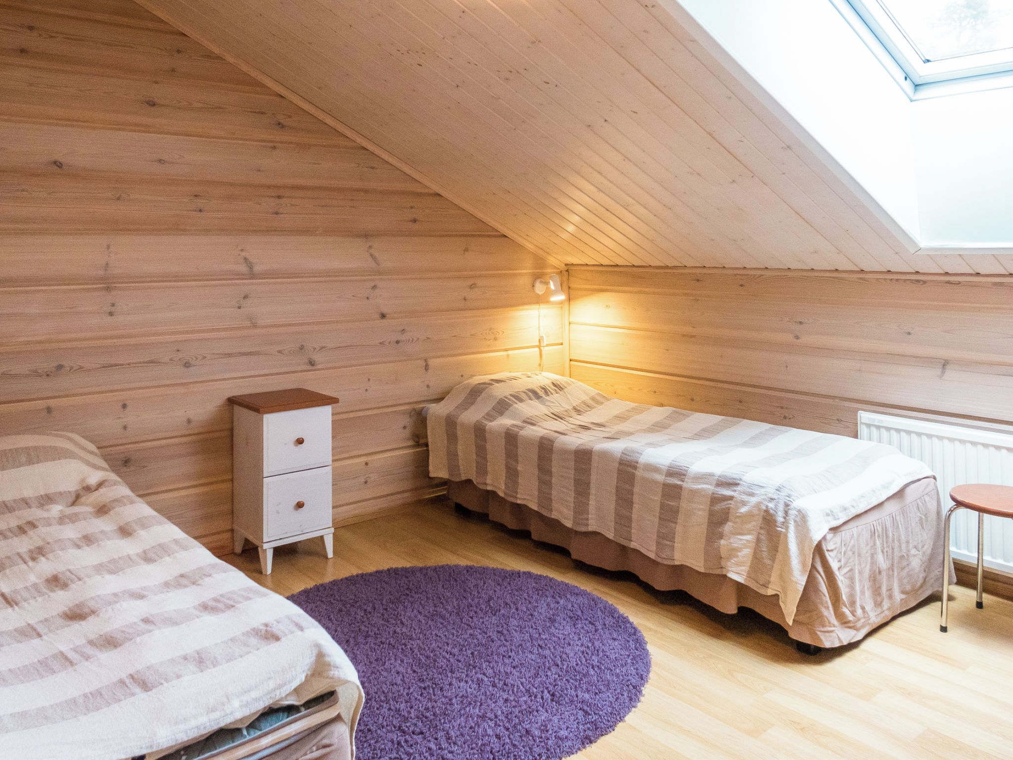 A small, cozy room with wooden walls and a sloped ceiling, featuring a bed, a nightstand, a dresser, and a window with a view.