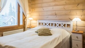 A neatly made bed with white bedding and a green pillow, flanked by two bedside tables with lamps, in a room with wooden panel walls and a window with curtains. - Klz 11. jade by Interhome (Sotkamo)