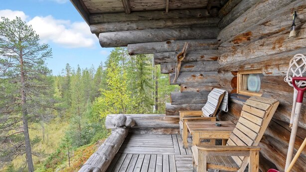 A log cabin with a wooden deck, two wooden chairs, a red shovel, and a broom leaning against the cabin wall.