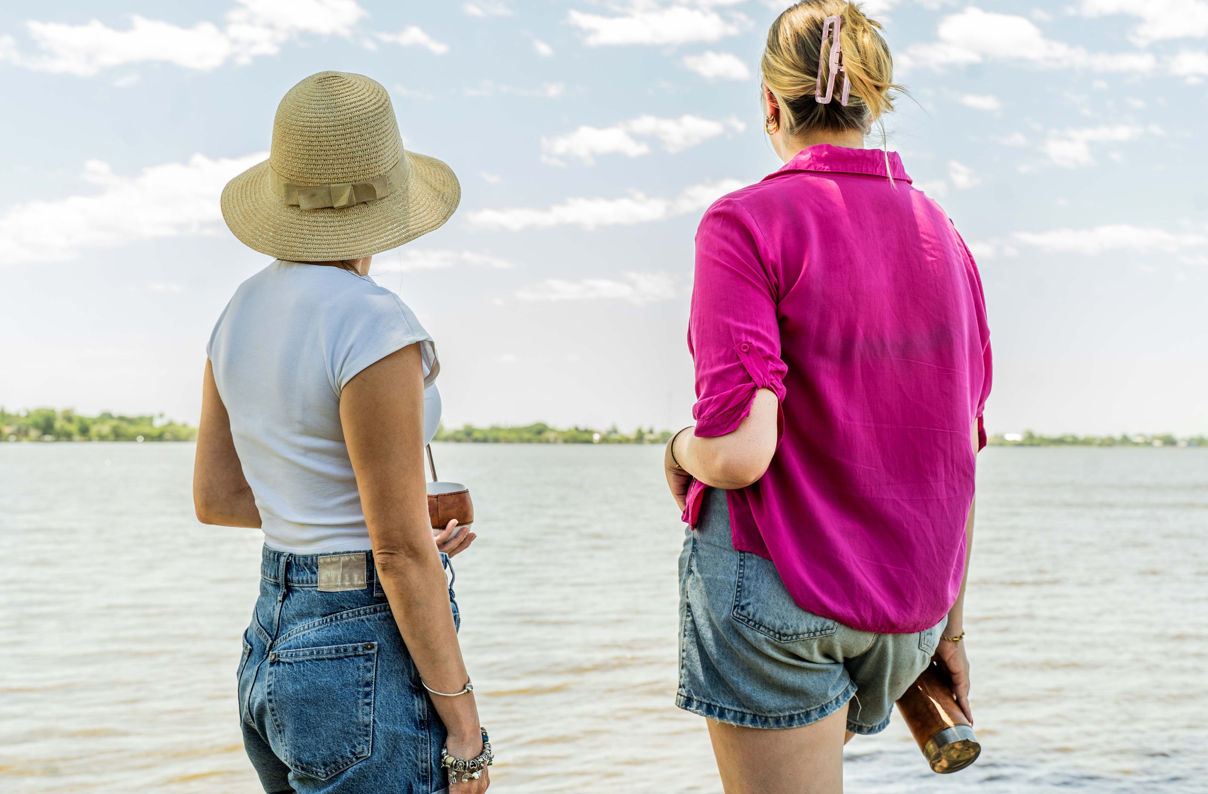 Two individuals standing by a body of water, one wearing a straw hat and denim shorts, the other in a pink shirt and denim shorts.