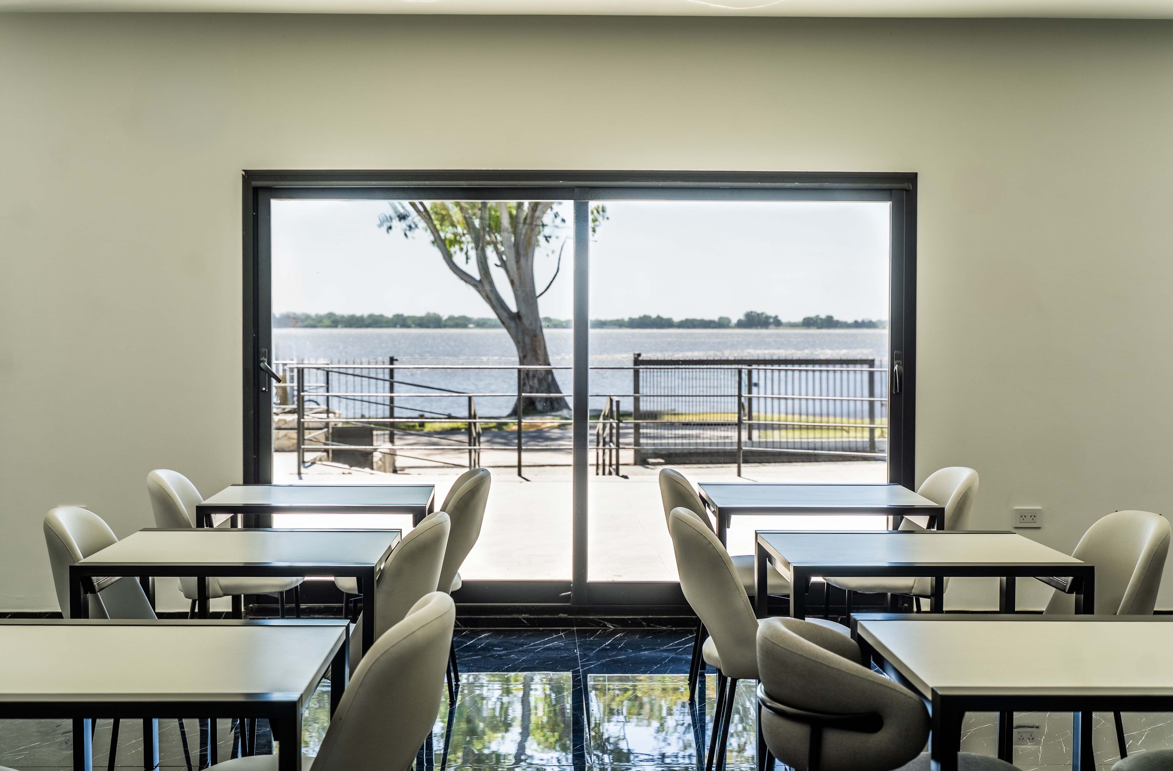 A room with a large window overlooking a body of water, a tree, and a railing. There are tables and chairs arranged for dining.
