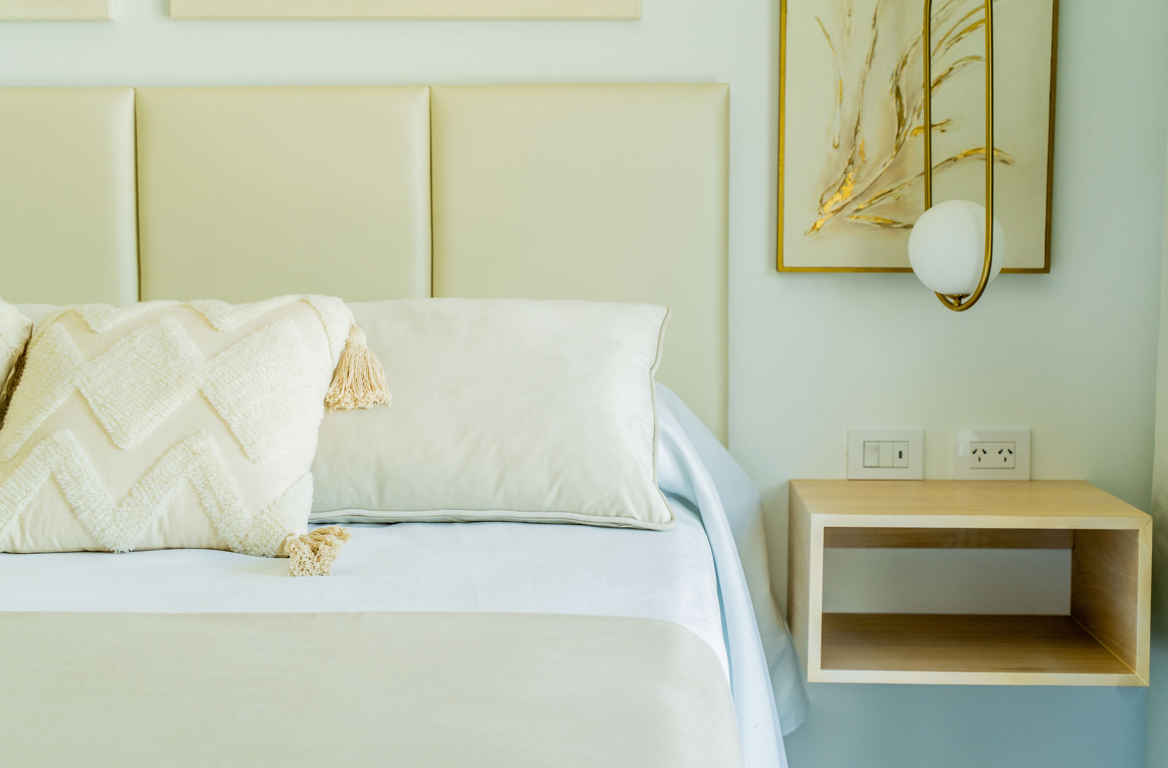 A bed with a white duvet and pillows, a bedside table with a lamp, and a wall-mounted power outlet.