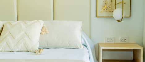 Una cama con edredón blanco y almohadas, una mesita de noche con lámpara y un enchufe en la pared.