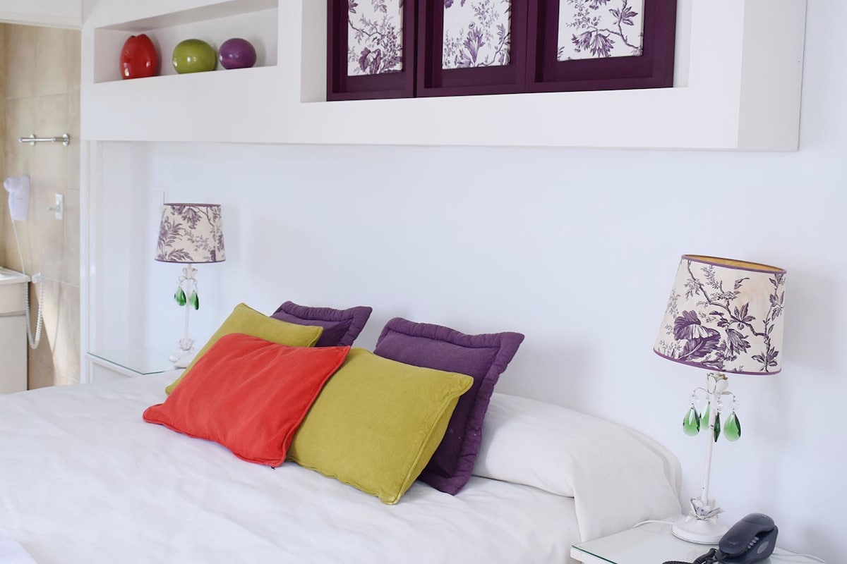A hotel room with a bed, a bedside lamp, a mirror, and a shelf with decorative items.