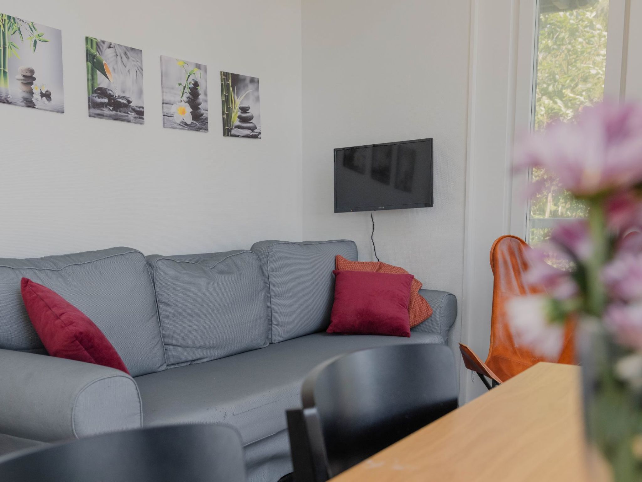 Moderni olohuone, jossa on harmaa sohva, punaiset ja oranssit koristetyynyt, seinälle asennettu taulutelevisio ja ikkuna, josta avautuu näkymä vehreään maisemaan.