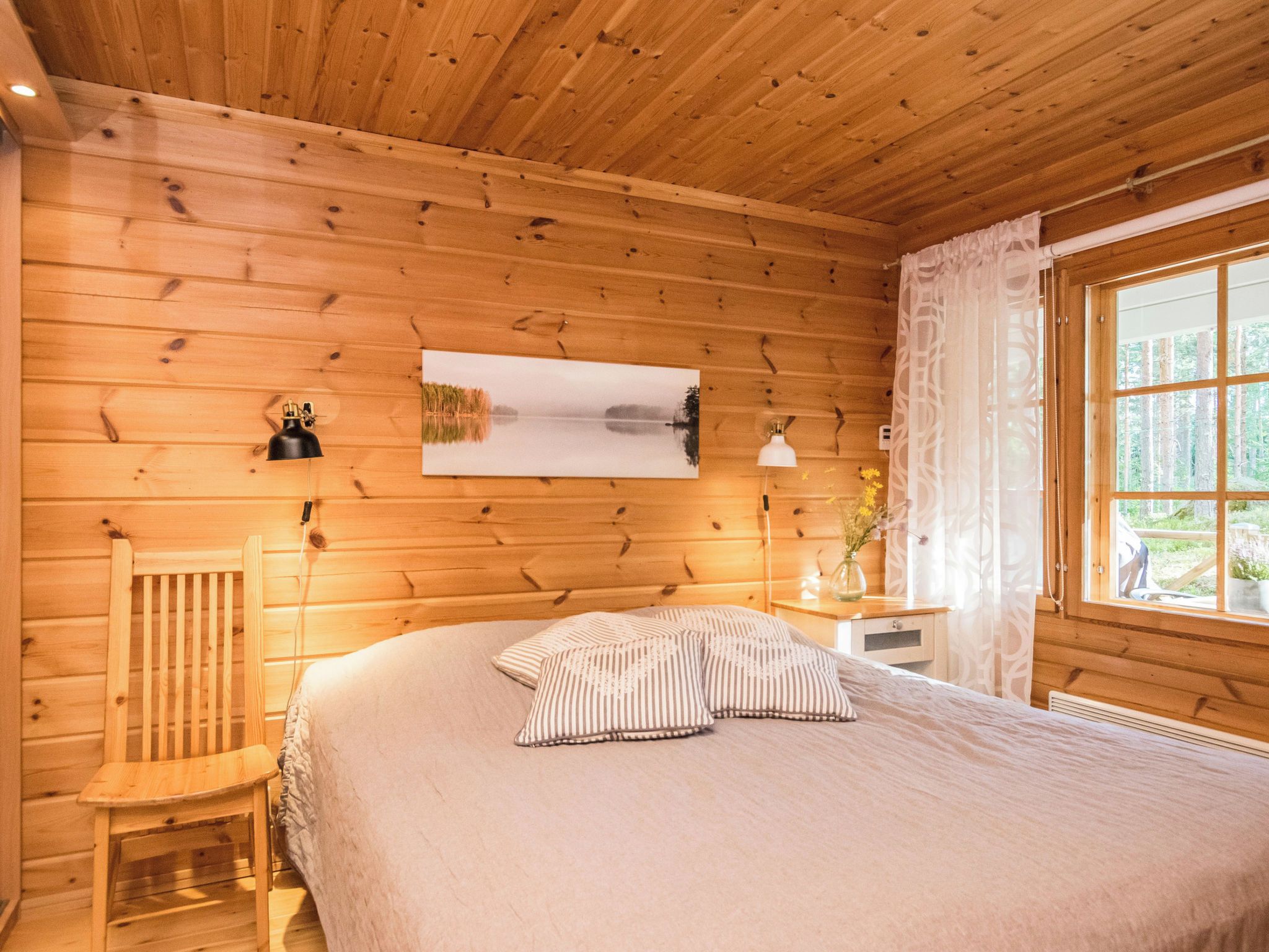 Ein Schlafzimmer einer Holzhütte mit einem Bett, einem Sessel, einem Nachttisch, einer Lampe, einem Fenster mit Vorhängen und einem Bild an der Wand.
