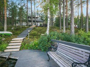 A wooden deck with a bench, surrounded by a forested area with a house in the background.