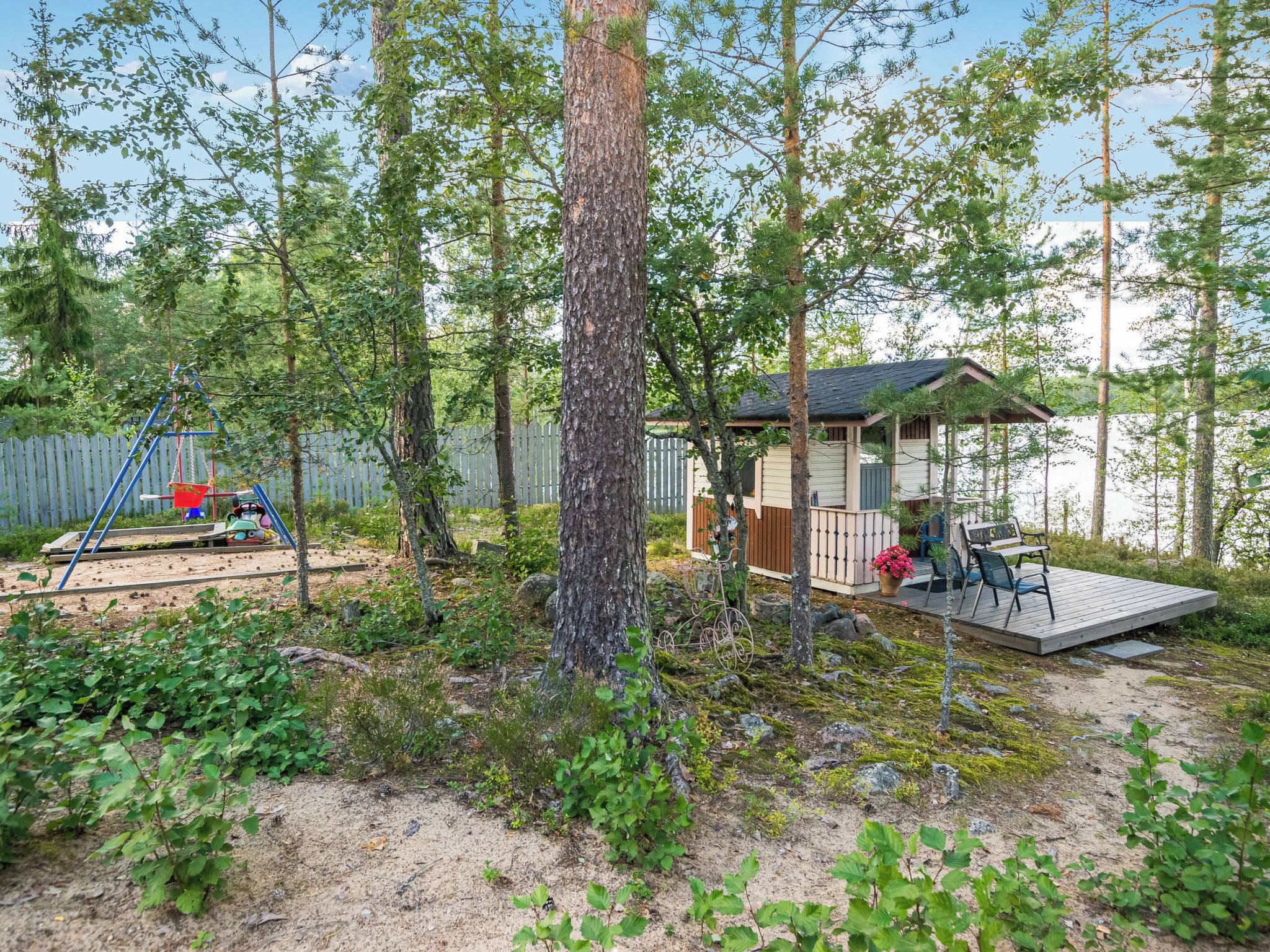 En træterrasse med udendørs møbler, et lille skur og et gyngestativ i et skovområde.