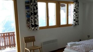 Une pièce comprenant une chaise en bois, un lit recouvert d’une couette à motifs floraux et une fenêtre avec des rideaux.
