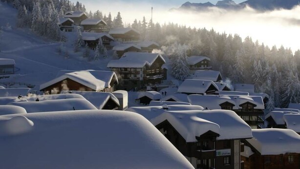 Un village enneigé aux bâtiments en bois, avec une chaîne de montagnes en arrière-plan et un ciel dégagé.