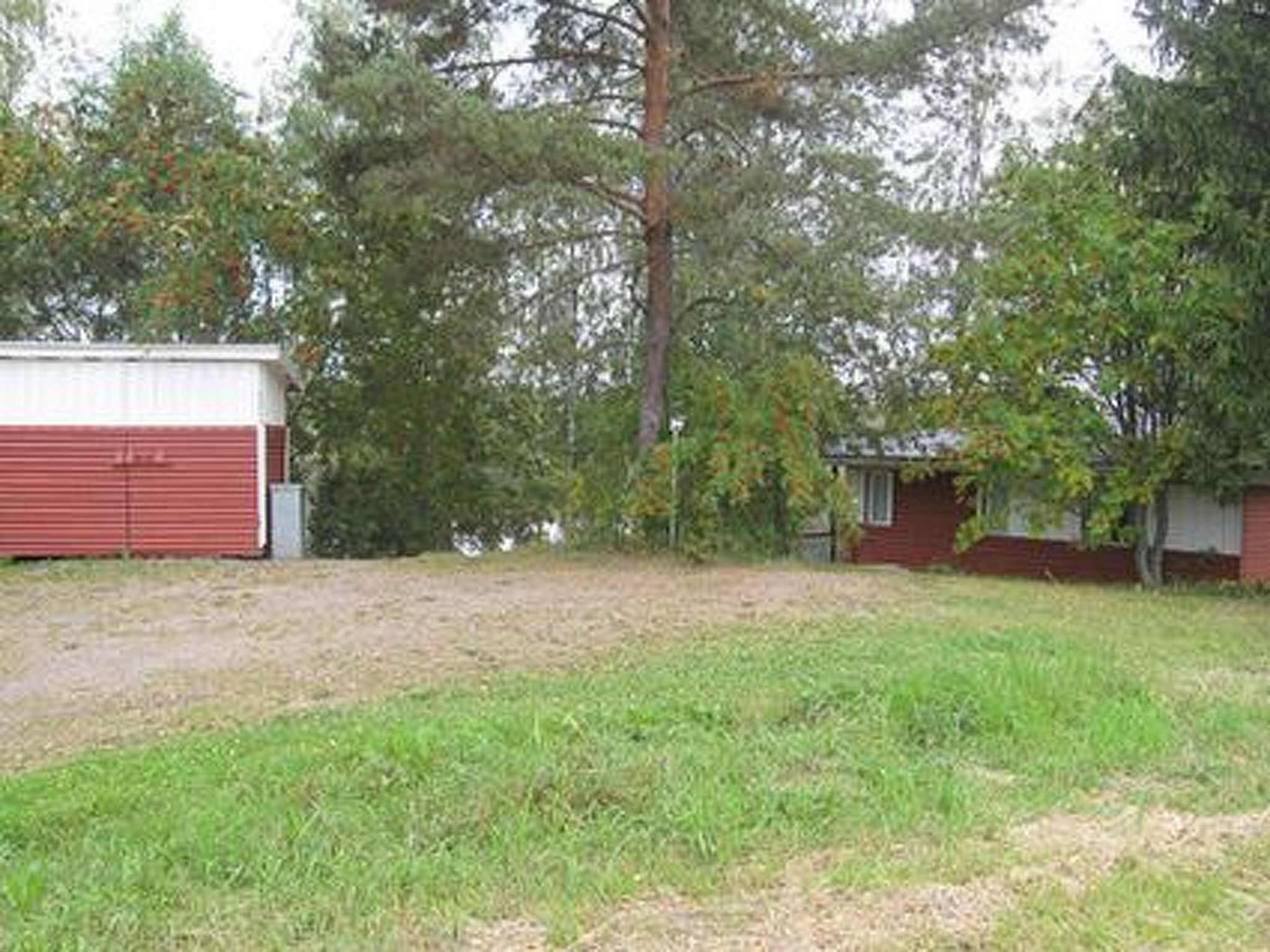 Ett rött garage och en liten byggnad med ett sluttande tak, omgivet av gräs och träd.