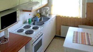 Een kleine keuken met een fornuis, spoelbak en een magnetron. Er is een tafel met een placemat en een raam met gordijnen.