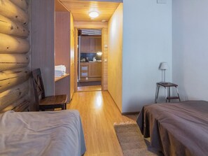 A narrow hallway leading to a kitchen area with wooden walls and a ceiling light. There are two beds with wooden frames and a small table with a lamp.