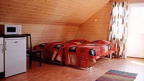 Une petite pièce chaleureuse aux murs en bois, avec un lit recouvert d’une couverture rouge, un micro-ondes posé sur une petite table et une fenêtre ornée de rideaux.