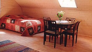 Une chambre mansardée chaleureuse avec un plafond en pente en bois, un lit rouge matelassé, une table à manger avec des chaises et un vase rempli de fleurs.