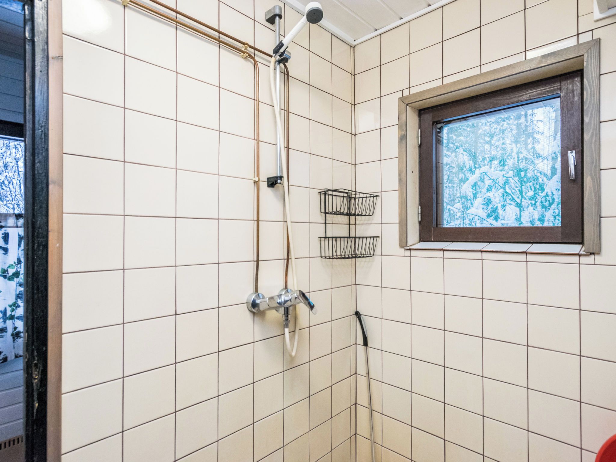 Kylpyhuoneessa on valkoisilla laatoilla päällystetyt seinät, käsisuihkulla varustettu suihku, ikkuna, josta avautuu näkymä puita kohti, ja pieni musta hylly.