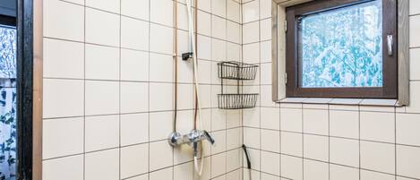 Ein Badezimmer mit weiß gefliesten Wänden, einer Dusche mit Duschbrause, einem Fenster mit Blick auf Bäume und einer kleinen, schwarzen Ablage.