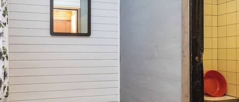 Un baño con pared blanca, un espejo, un banco y un cubo de basura rojo.