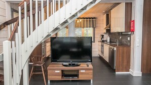 A modern living room with a flat-screen TV on a wooden stand, a staircase with white railings, and a kitchen area with wooden cabinets and a red sign. - Rukanloisto b by Interhome (Kuusamo)