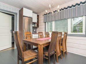 A dining area with a wooden table and chairs, a kitchen with a stainless steel refrigerator, and a window with checkered curtains.