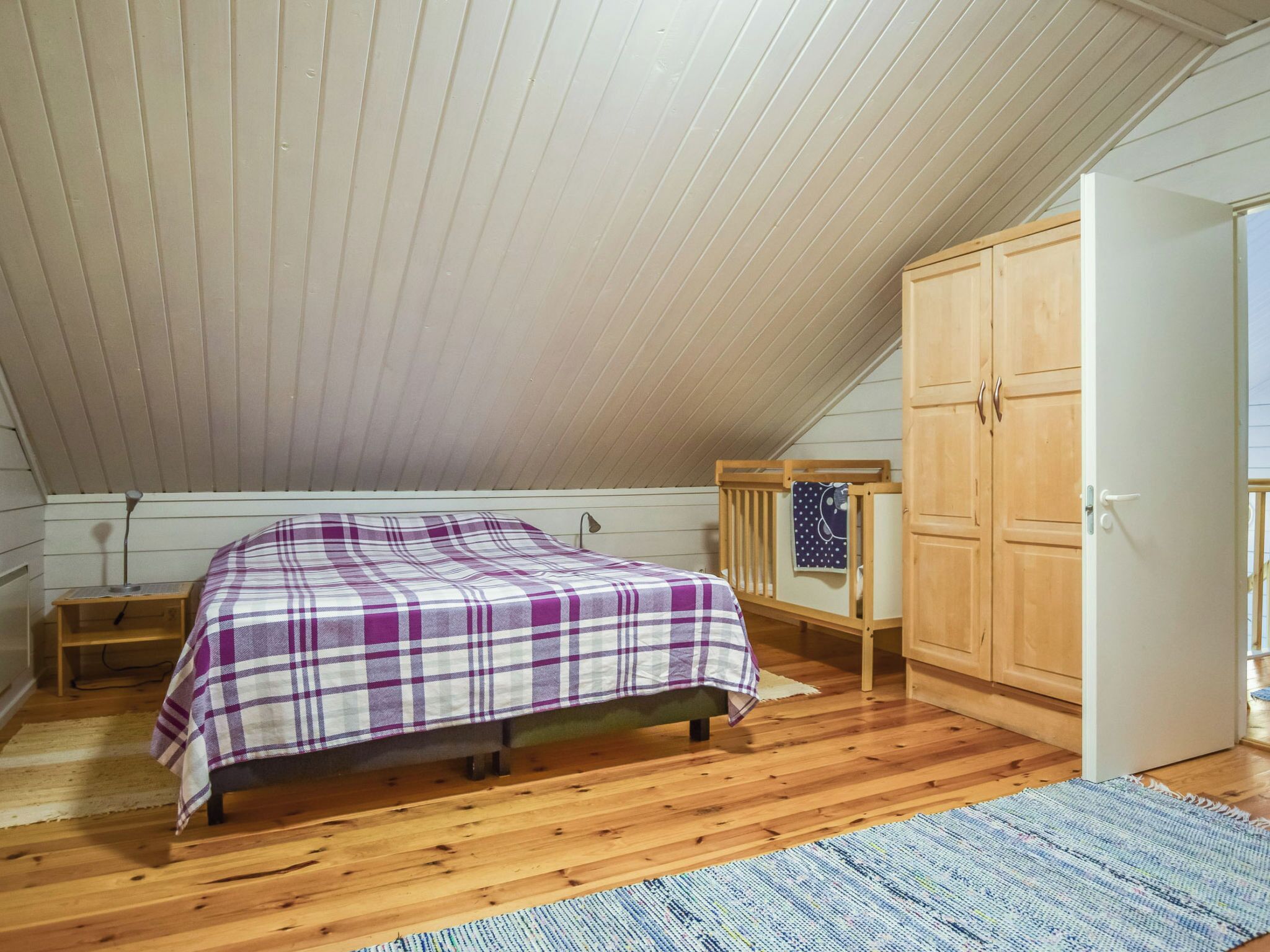 A bedroom with a wooden floor, a bed with a plaid bedspread, a nightstand, a wooden crib, and a wardrobe.