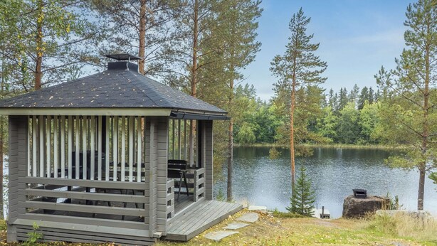 Un quiosco de madera con techo, banco y una pequeña mesa, situado junto a un lago rodeado de árboles.